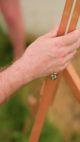 un primer plano de la mano de una persona, con una camisa a cuadros, ajustando un trípode de madera en un campo cubierto de hierba. el área circundante está borrosa, con la pierna de una mujer visible en el fondo
