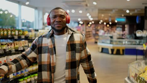 Portrait-of-a-happy-man-with-Black-skin-color-in-a-plaid-shirt-and-red-wireless-headphones-dancing-while-in-a-large-modern-grocery-store.-Happy-man-dancing-during-his-shopping-trip-and-shopping-at-the-grocery-store