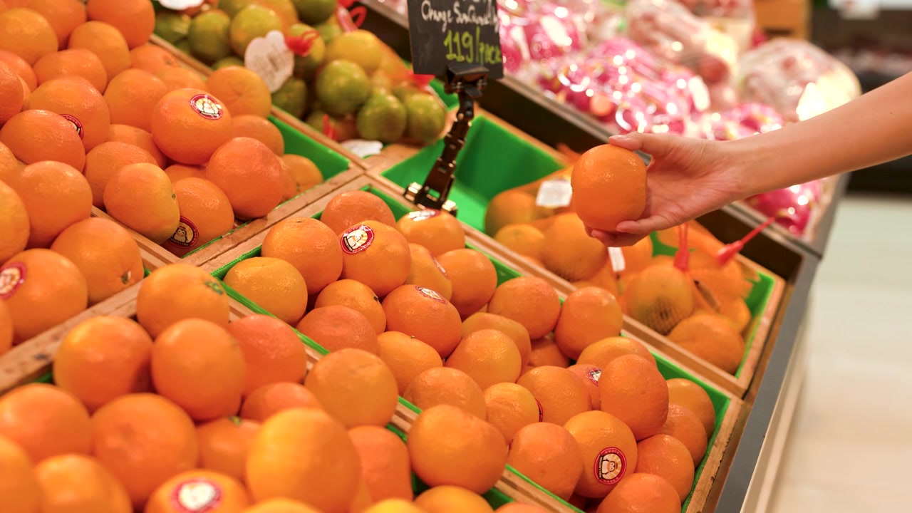 Close-up shot of a hand picking up and inspecting an orange in a grocery store.