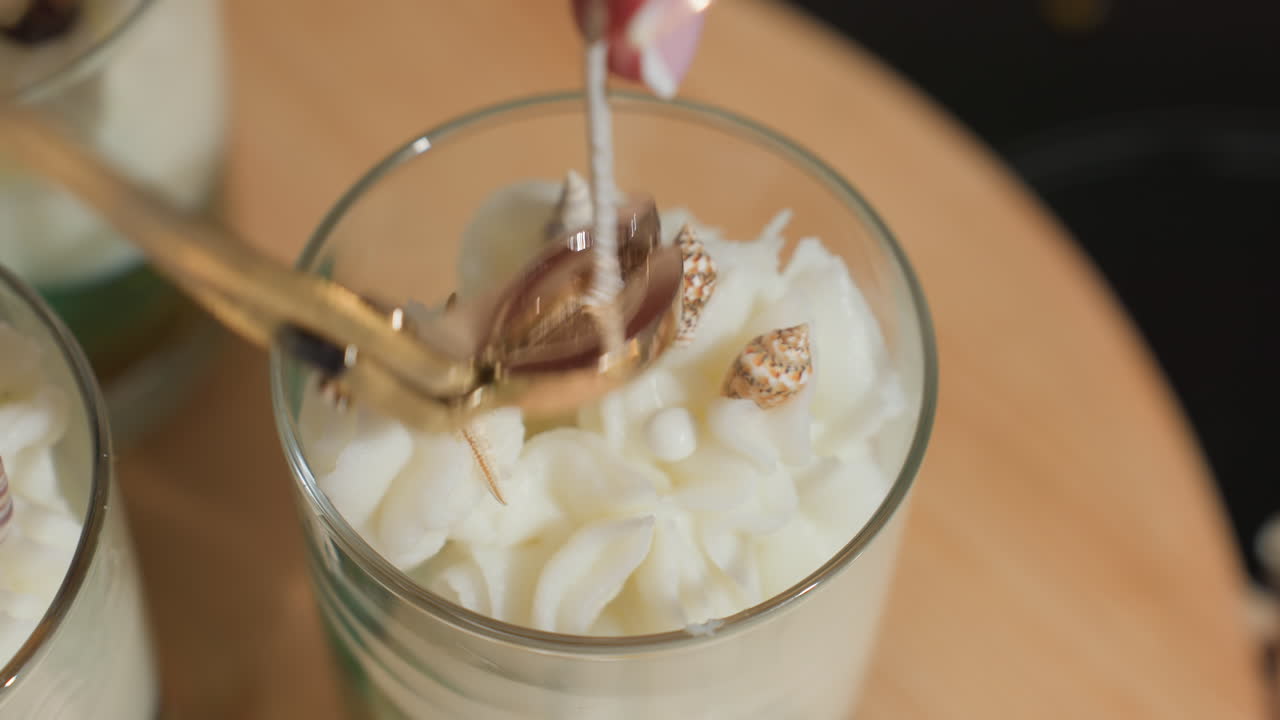 Closeup view of baker delicately trimming candle wick using gold trimmer on cake-styled wax creation adorned with whipped texture and small seashell decorations in glass jar, set on wooden surface