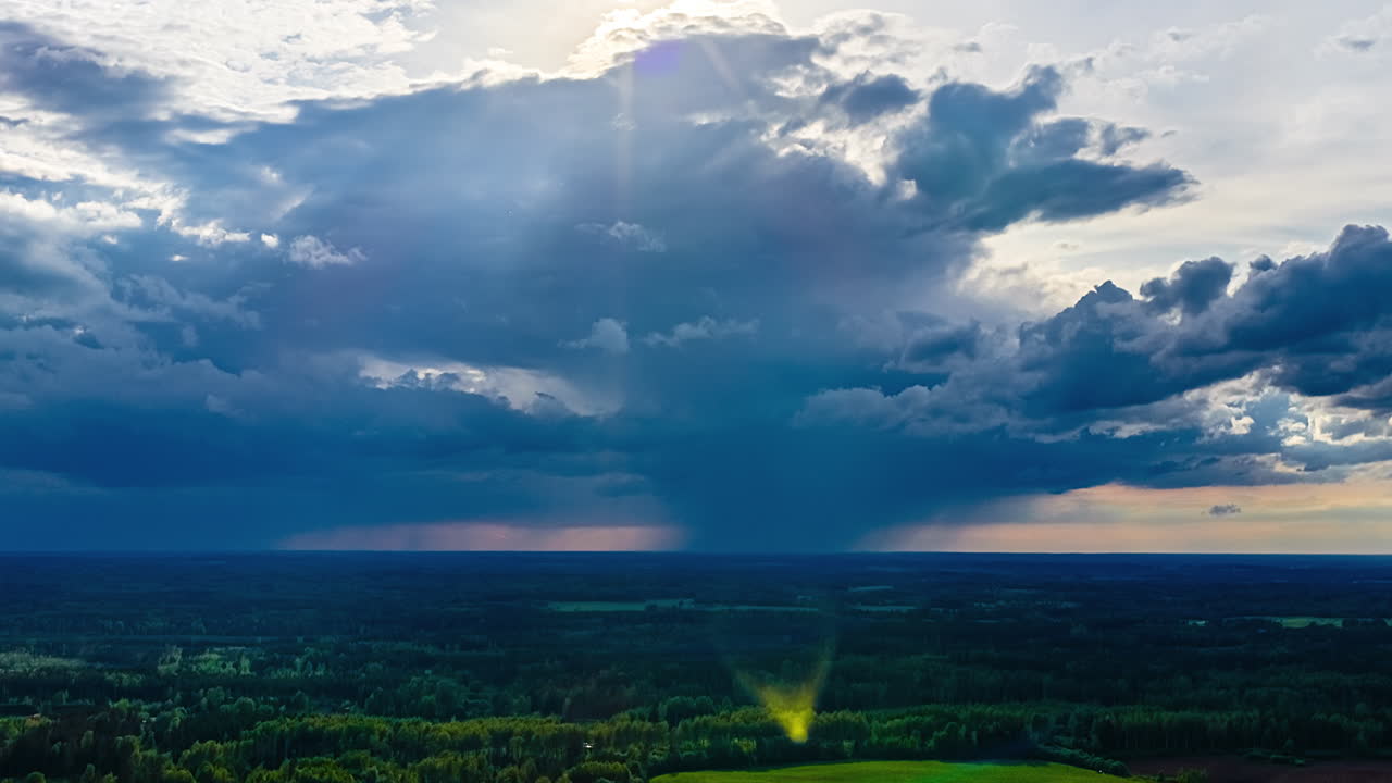 An epic aerial timelapse captures dramatic storm clouds, a rain shower, and beautiful sunbeams (god rays) shining down like a spotlight on a vast green forest