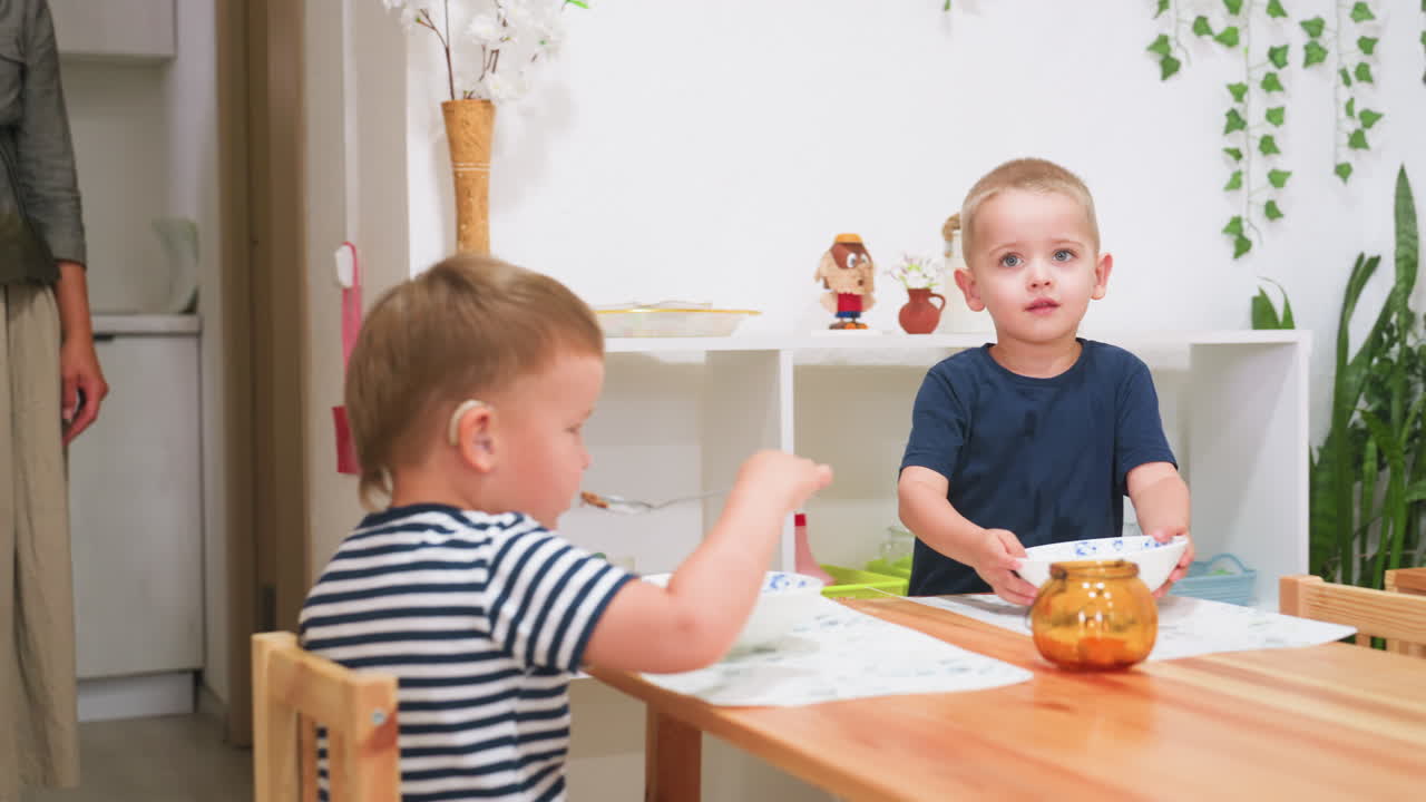 Toddler in kindergarten holding bowl walking to drop food on table while teacher places plate on dining surface, another child with hearing aid eating in background