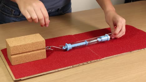 Hands use a spring scale to measure friction on wooden blocks over sandpaper. Close-up, well-lit, educational setup