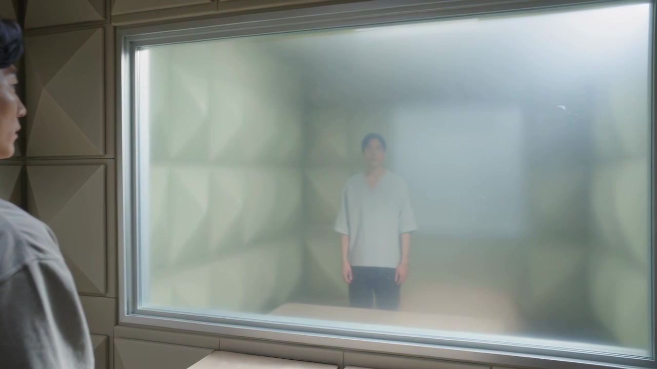 La telecamera si sposta rivelando un uomo in piedi all'interno di una cella, mentre un osservatore lo guarda attraverso una finestra smerigliata per una revisione.
