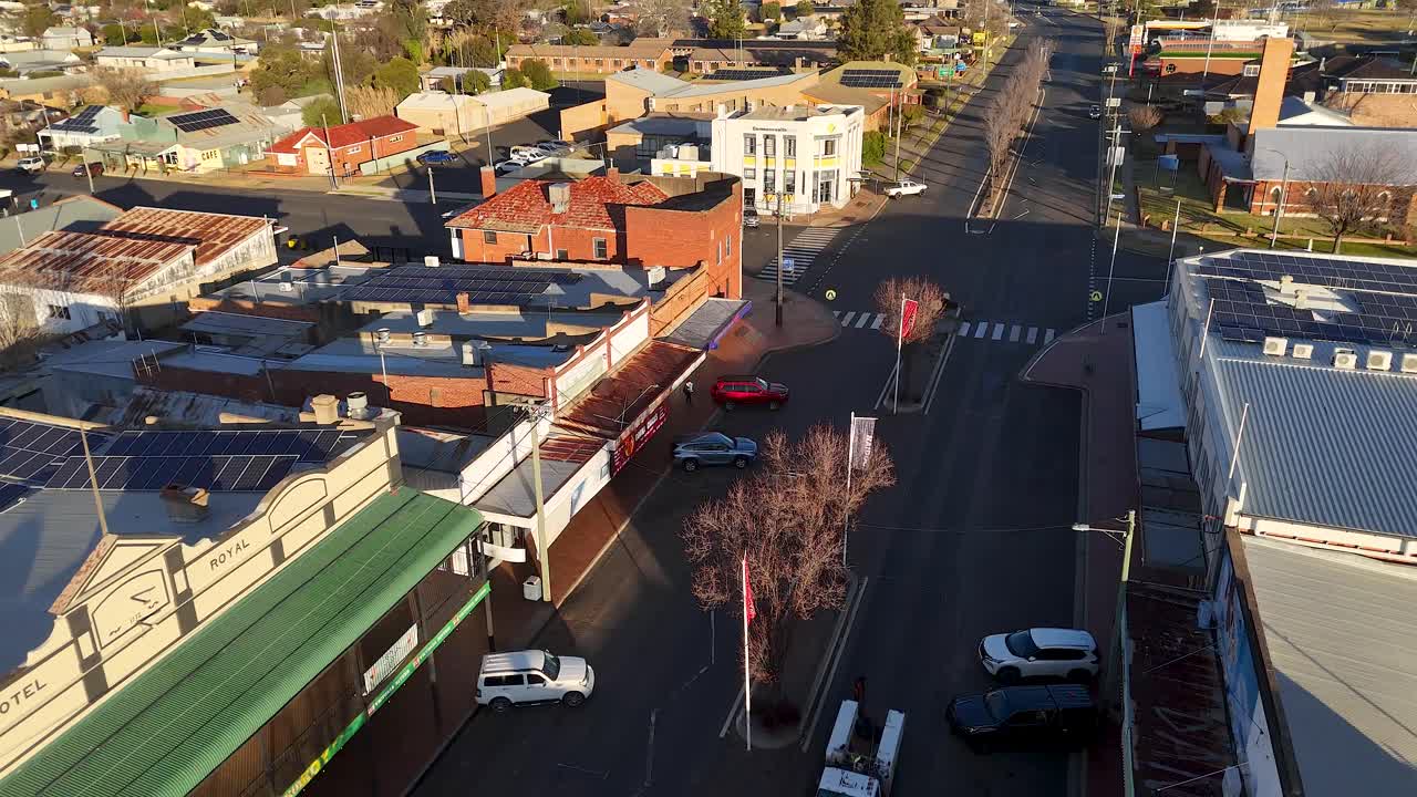 Overhead drone footage captures vehicles moving along a quiet urban street lined with shops and residential buildings in late afternoon sunlight