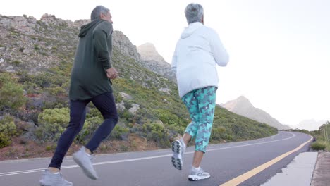 Happy-senior-biracial-couple-running-on-road-in-mountains