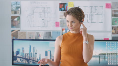Female-Architect-Standing-In-Office-Talking-To-Client-Using-Wireless-Earphones