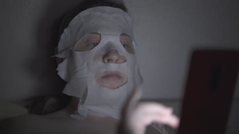 young-woman-in-regenerating-sheet-mask-lies-in-dark-room