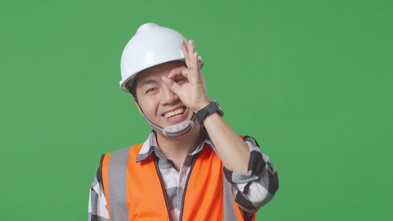 Close Up Of Asian Male Engineer With Safety Helmet Showing Ok Hand Sign Over Eye And Smiling To Camera While Standing In The Green Screen Background Studio