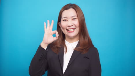 beautiful businesswoman in showing ok hand gesture smiling standing on blue background looking at camera