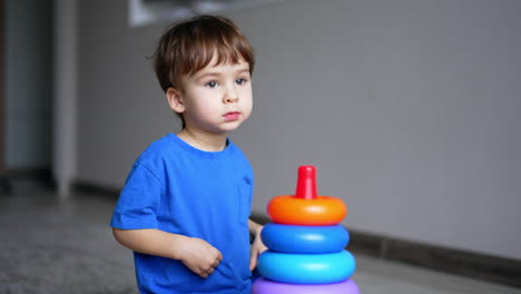 caucasian toddler in blue t-shirt plays with toy pyramid. kid looks ahead and points at something with his finger.