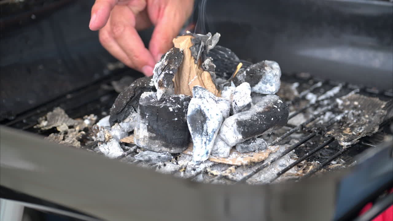 Slow motion close up of a male hand firing a piece of cardboard using a match to start a grill with burnt charcoal