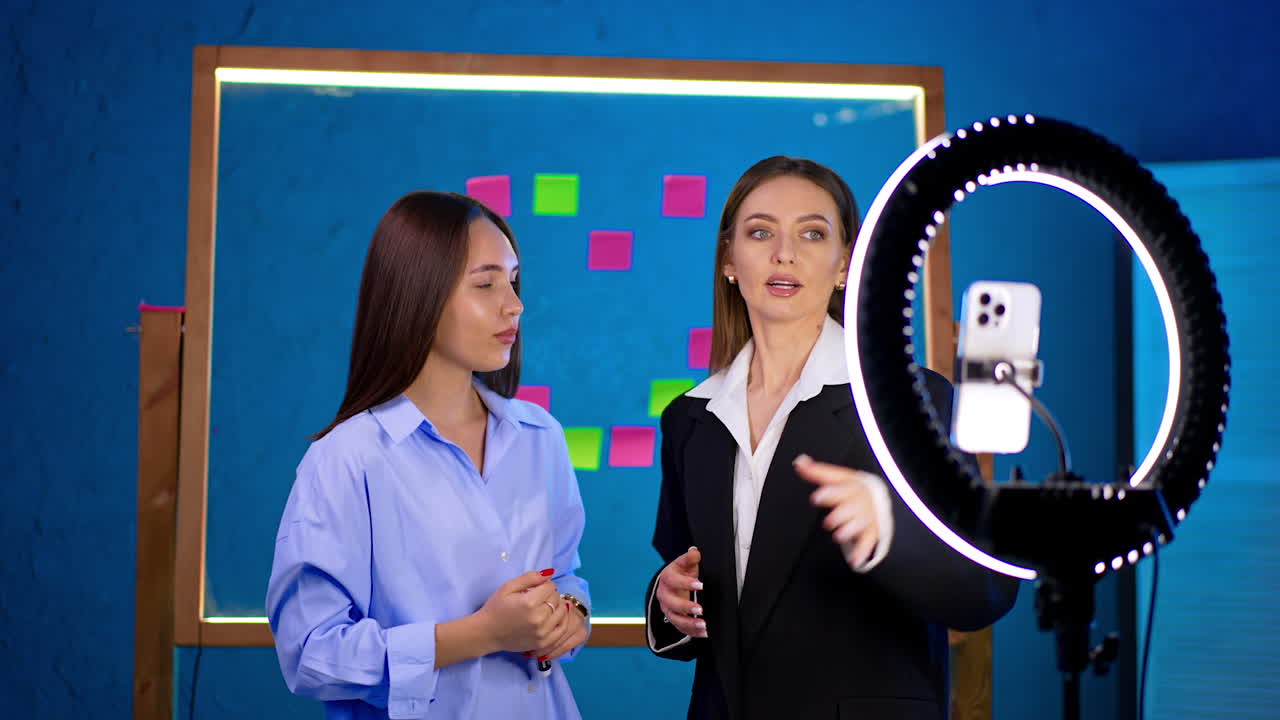 Two long-haired brunettes stand at the phone with ring light. Women talk, discuss, prepare for blog footage.