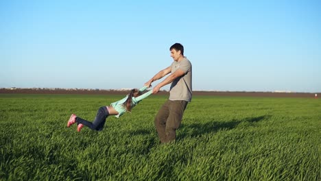 young father in casual spinning his daughter in circles, playing, having fun in nature on a bright day. parenthood. beautiful skyline, green field