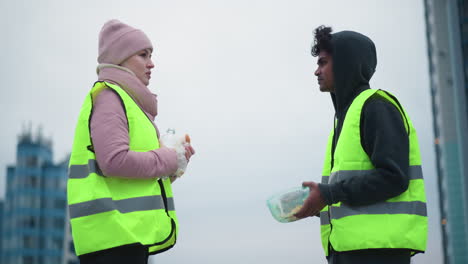 Two workers in neon yellow reflective vests take break outdoors in cold weather, smiling and talking while eating food from plastic container and drinking water, dressed warmly in hoodies, jackets
