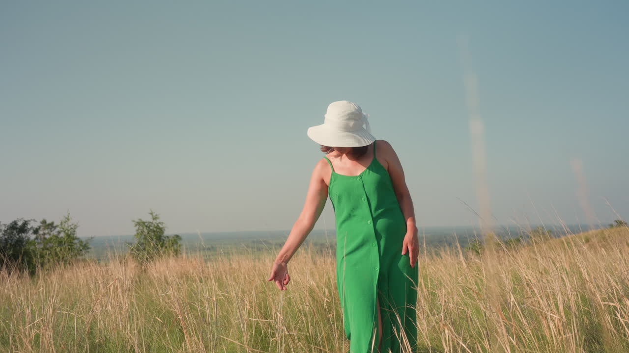 woman in green dress and white hat gracefully bends in tall golden grass, gently pulling out strand while looking down with soft smile under sunny sky