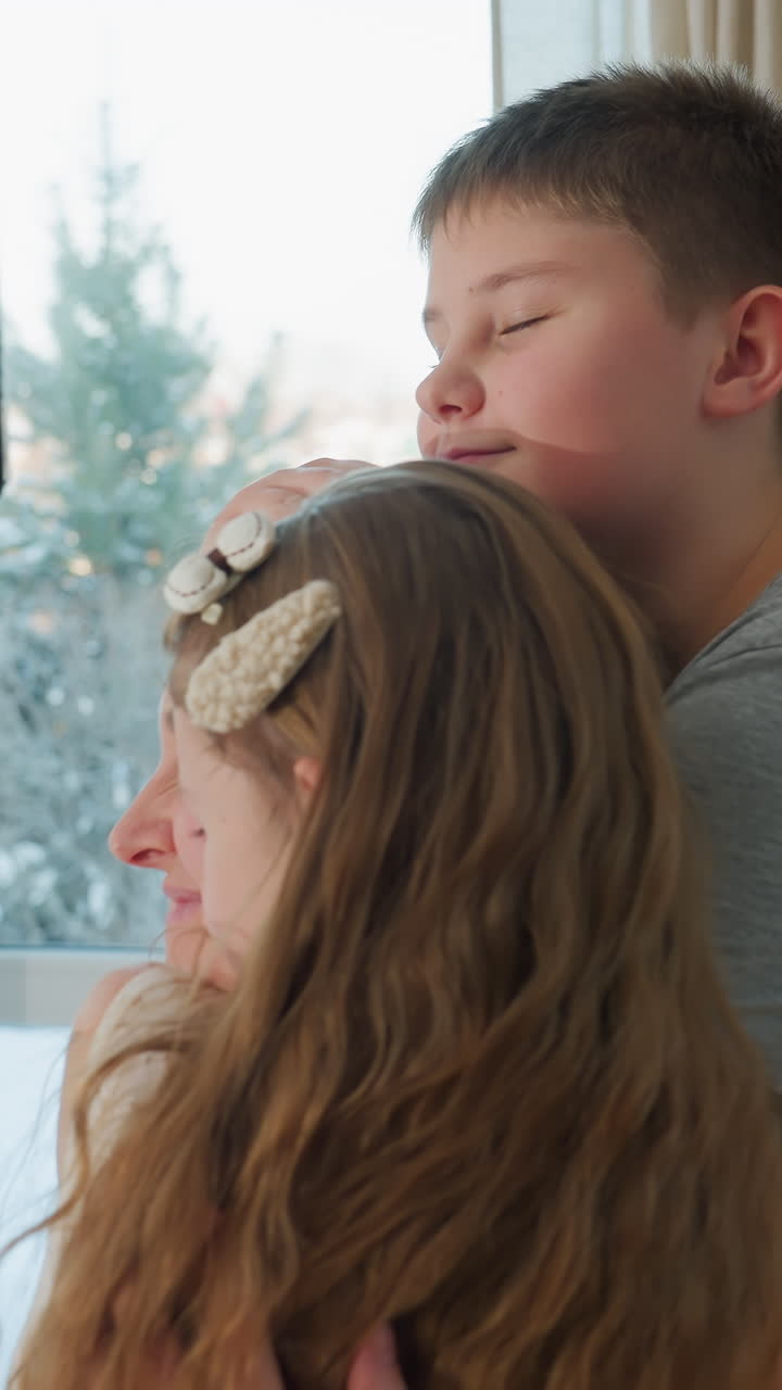 Madre e hijos se abrazan junto a la ventana, tierno abrazo entre hermanos con toques afectuosos, madre sentada a la mesa de cristal, luz invernal suave a través de las cortinas, calidez doméstica y emotivo reencuentro