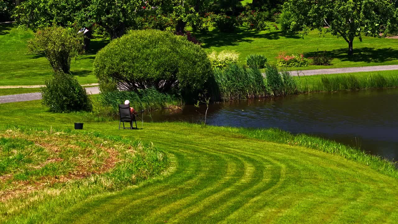 A person sits alone on a chair by a calm lake, surrounded by green fields and tall grass