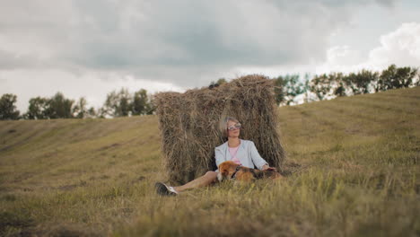 young woman wearing sunglasses and casual outfit sits against hay bale, gently petting dog while enjoying peaceful farmland, rolling hills, golden grass, and a cloudy sky