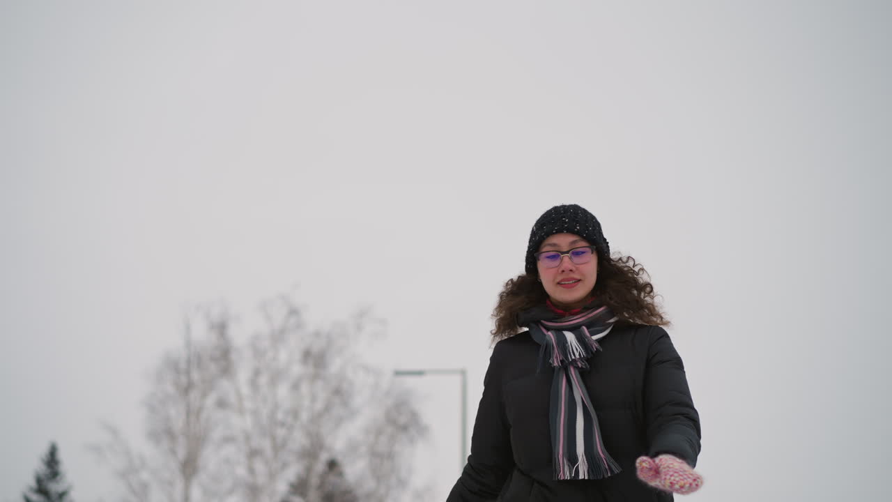 Athletic girl skating outdoors in winter snow wearing black coat, knit hat, colorful scarf and glasses, balancing with arms outstretched, enjoying cold weather activity with natural trees and cloudy sky