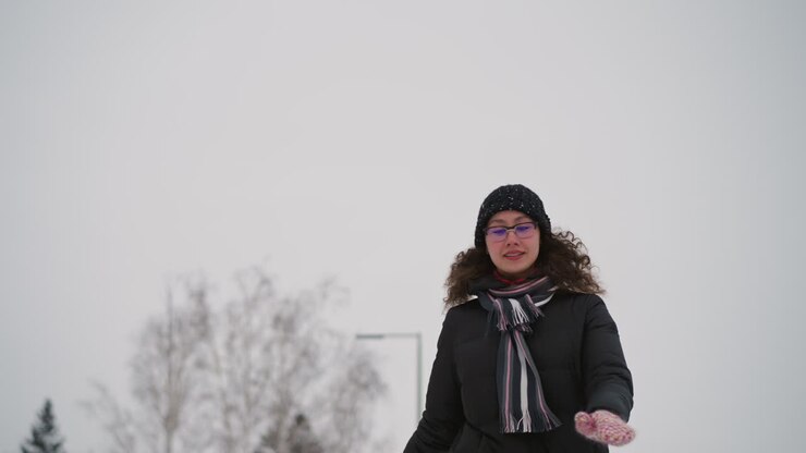 Athletic girl skating outdoors in winter snow wearing black coat, knit hat, colorful scarf and glasses, balancing with arms outstretched, enjoying cold weather activity with natural trees and cloudy sky