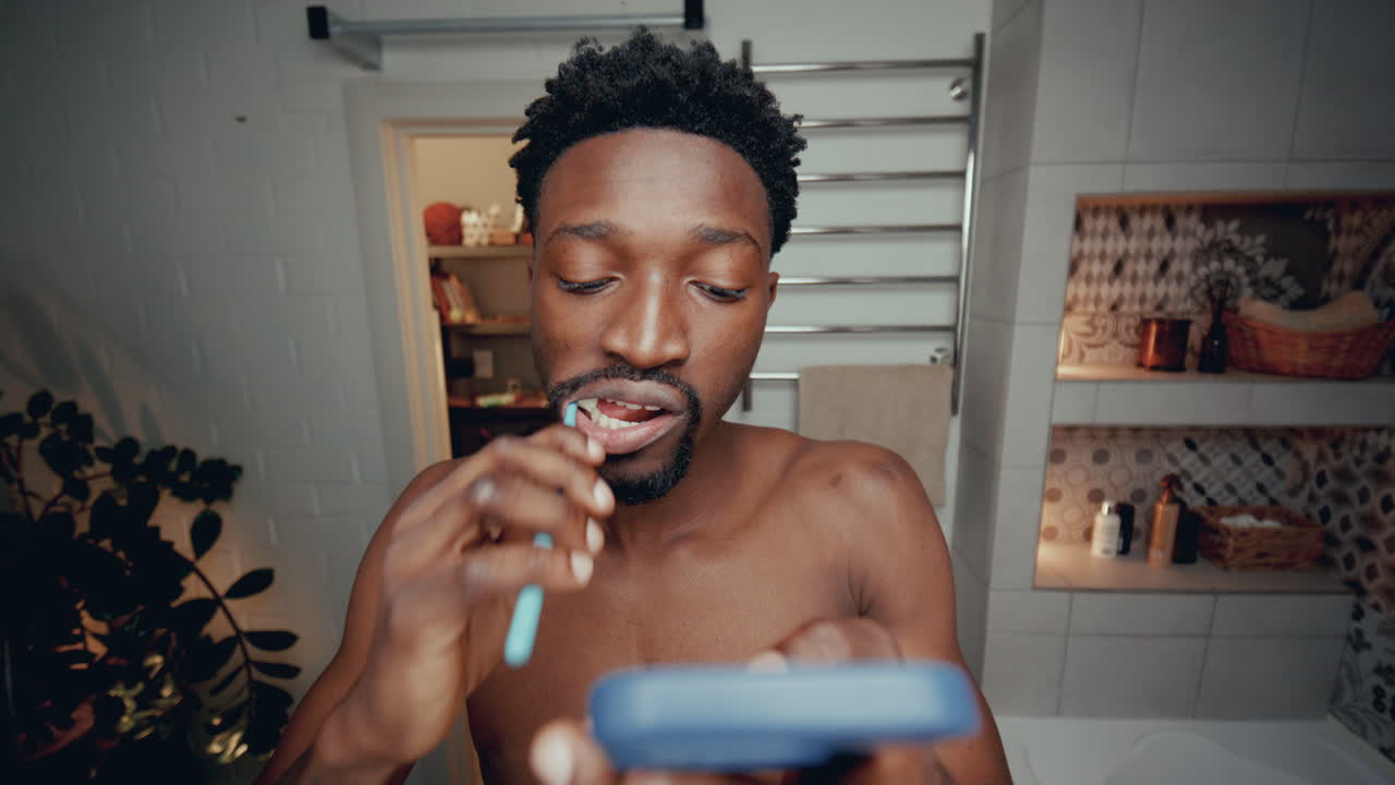 joven afroamericano cepillándose los dientes y usando el teléfono en el baño