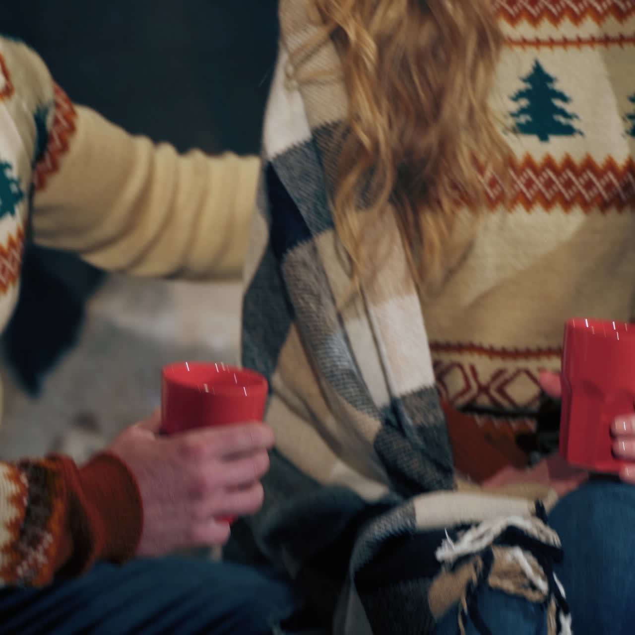 Young couple at Christmas time at home. Man and woman in Christmas sweaters sitting near fireplace with red cups. Romantic atmosphere of couple in love.