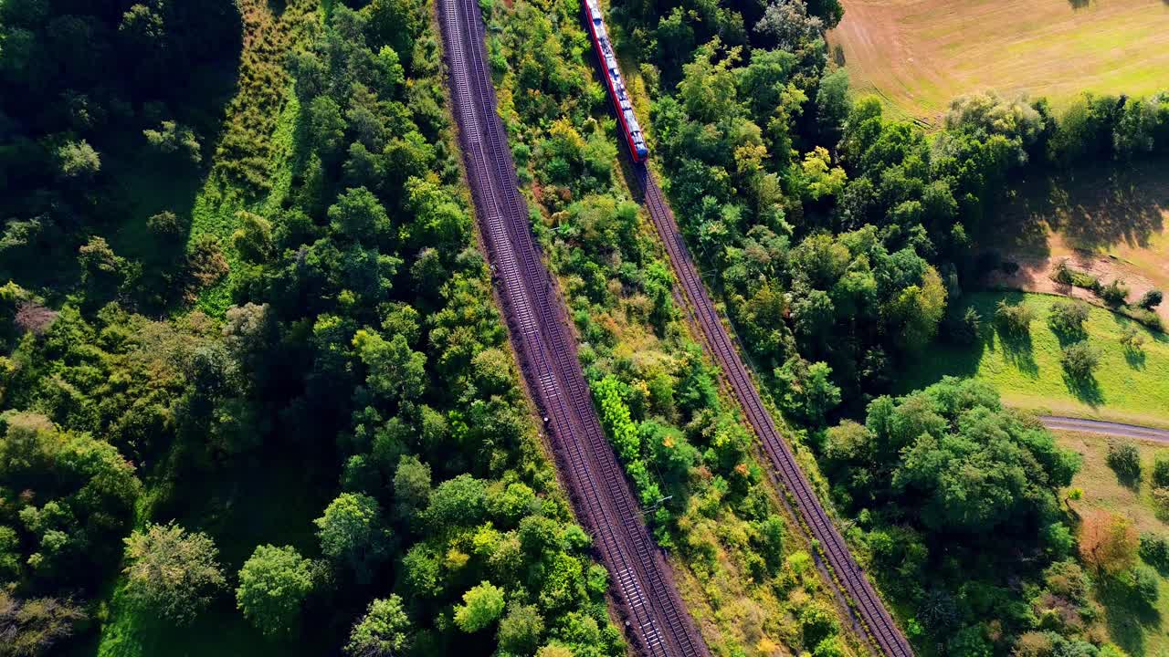 Aerial drone view of passenger train traveling through green forest along railway tracks with farmland nearby