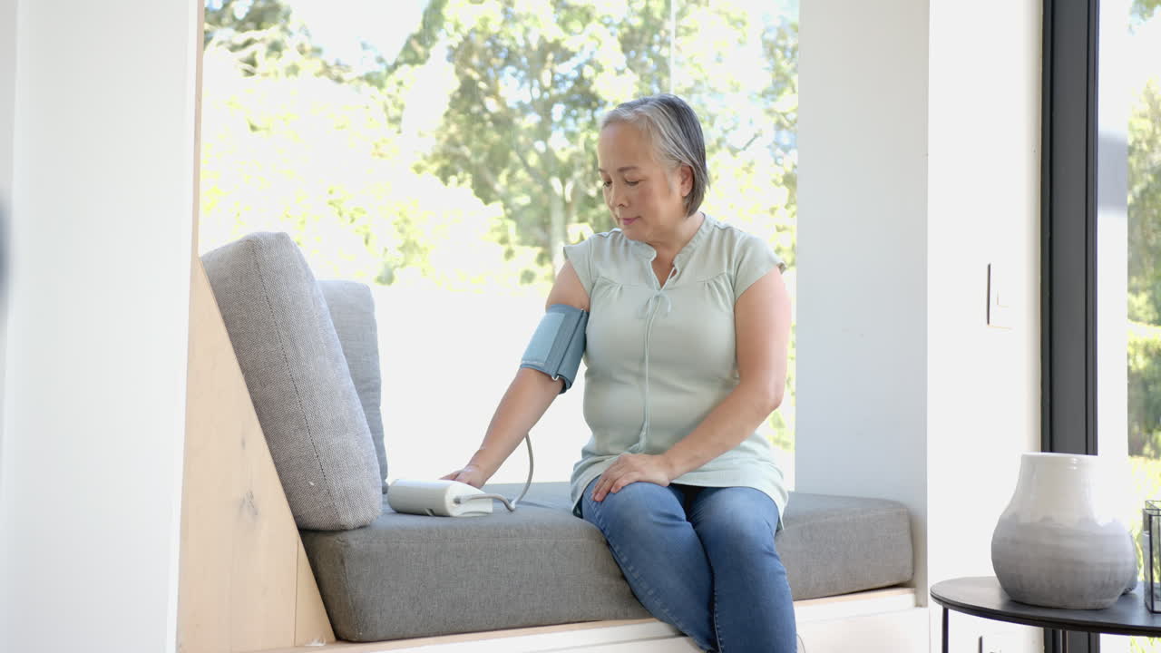 Measuring blood pressure, senior asian woman sitting on couch at home, focusing on health