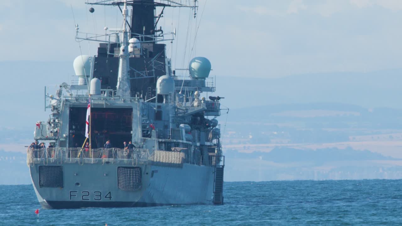 A large military vessel moves steadily forward on calm blue water, viewed from directly behind under clear daylight with distant coastline visible