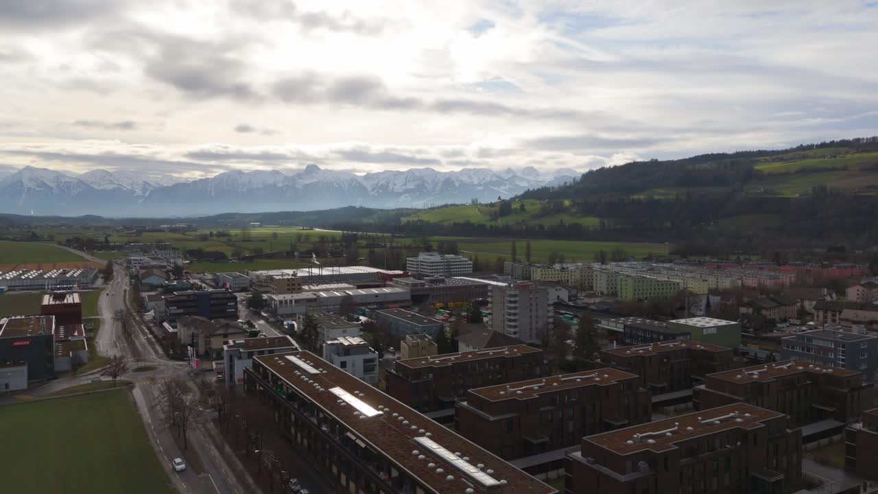 ciudad suiza con edificios en primer plano y montañas cubiertas de nieve en la distancia, cielos nublados por encima, vista aérea