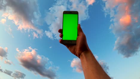 man's hand holds mobile smartphone with green screen on cloudy sky background