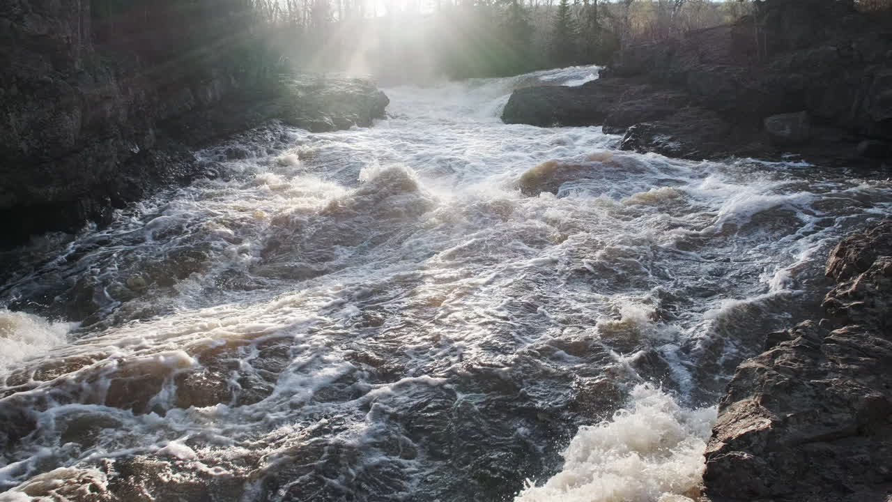 Slow pan and lens flare, raging rocky river rapids with water spray at sunset