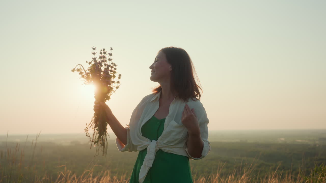 Sunlit woman in green skirt holds wildflower bouquet while turning joyfully on grassy river bank under golden sunset glow over river valley with gentle breeze and radiant backlight