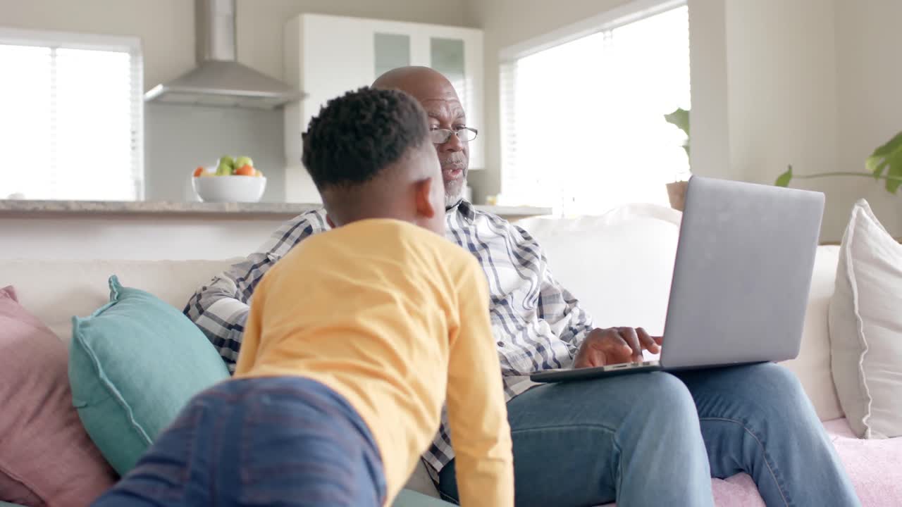 African american grandfather with grandson using laptop on couch at home, slow motion