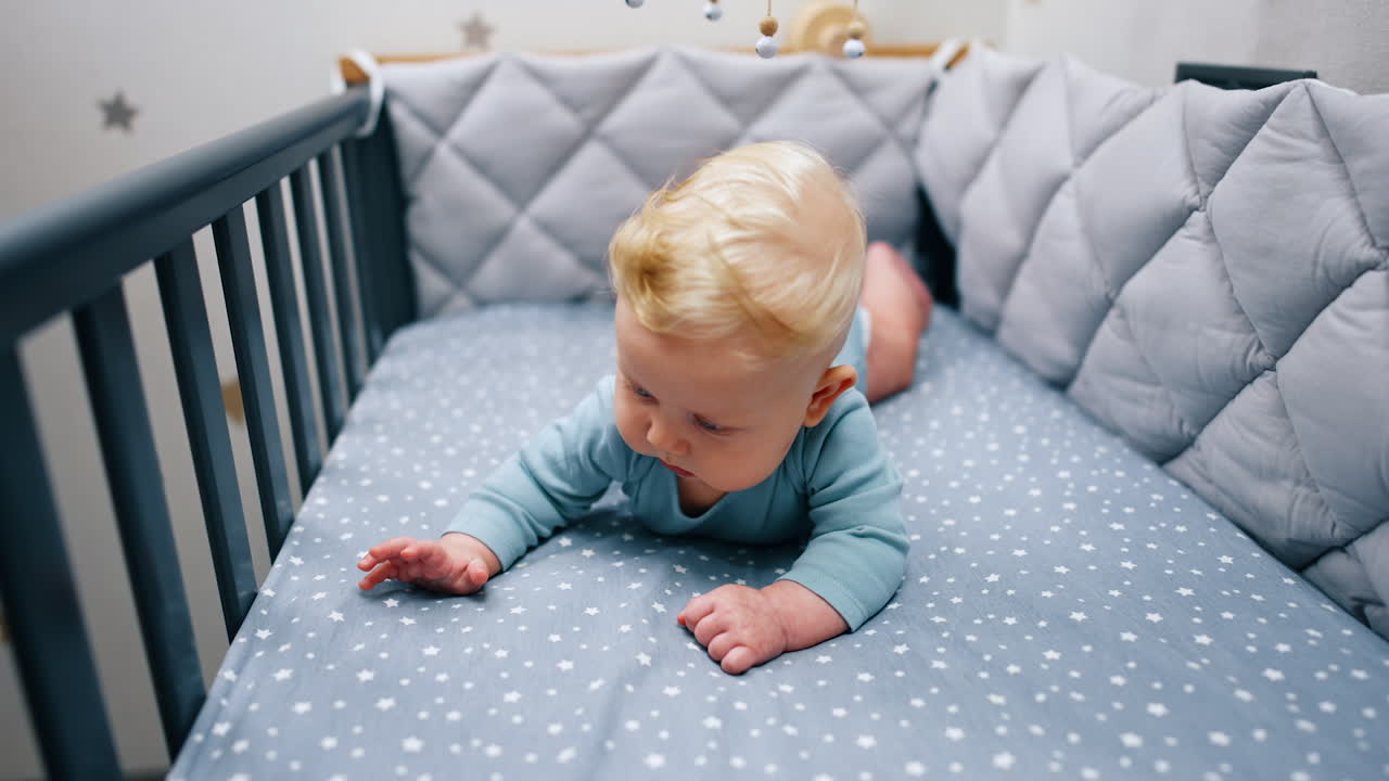 Little baby boy with big blue eyes lies on the belly. Cute infant wearing blue bodysuit in the cot.