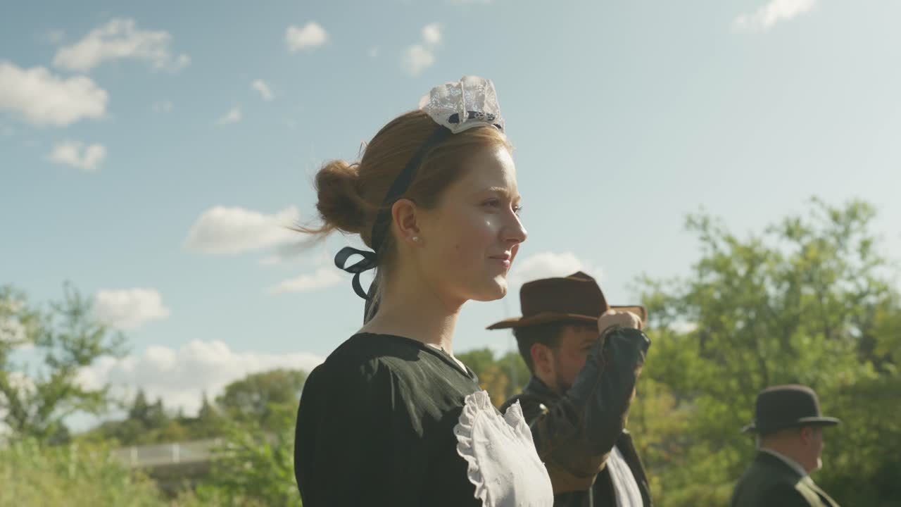 A Caucasian woman wearing an old maid dress stands outdoors in a grassy forest area, smiling lightly as the wind blows