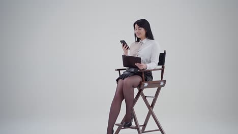 Attractive smiling woman holding phone and folder. Beautiful secretary sitting in the chair and looking on the phone in the studio. Slow motion.