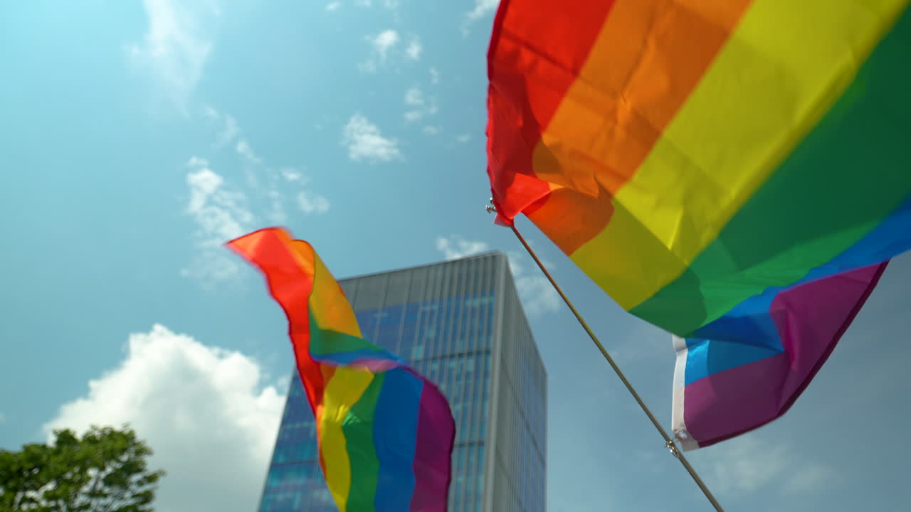 Flags in LGBT colors flutter in the wind, mounted on wooden and plastic poles - in the background, a modern glass building