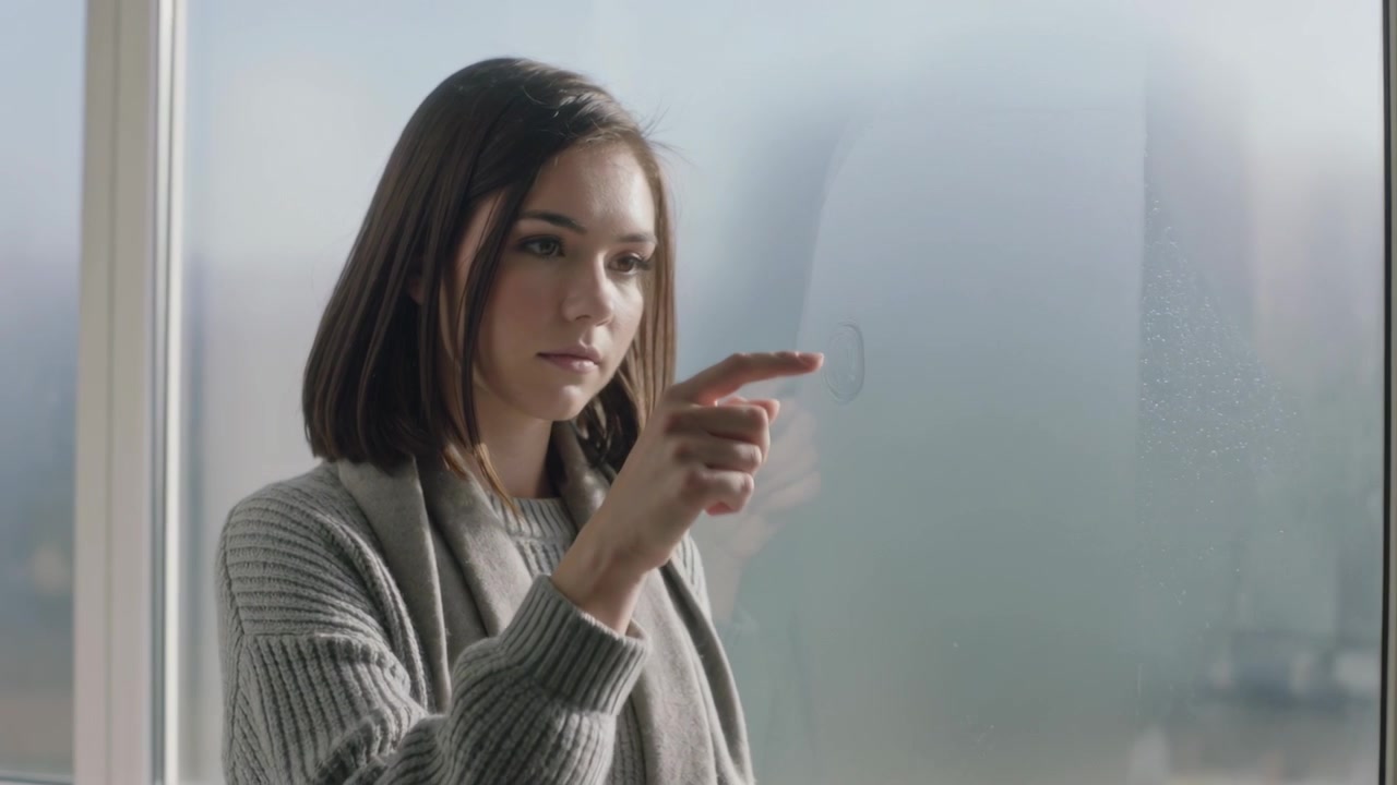 Mujer levantando el dedo, trazando con la yema en una ventana empañada para dejar una marca, con suéter gris y bufanda.