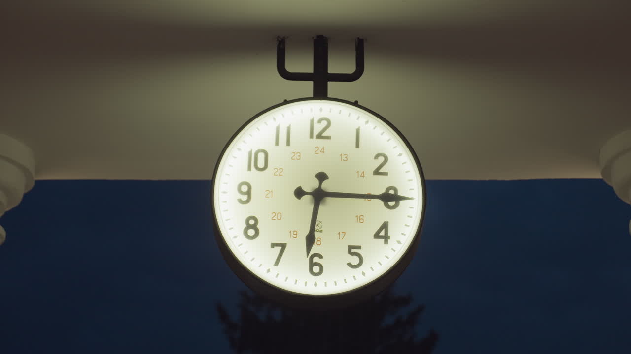 Close up of glowing round clock hanging on metal fork-like bracket under roof structure, surrounded by darkness of evening sky, casting soft ambient light over architectural elements of building top