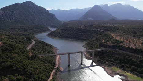 Vista-Aérea-De-La-Carretera-Strada-Provinciale-38-Que-Cruza-El-Río-Lago-Del-Cedrino-Con-Paisaje-Montañoso-Al-Fondo