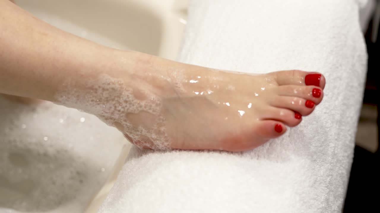 A woman takes her foot out of a bath with soapy water and places it on a pedicure stand, red gel polish pedicure, preparation for pedicure and softening the skin