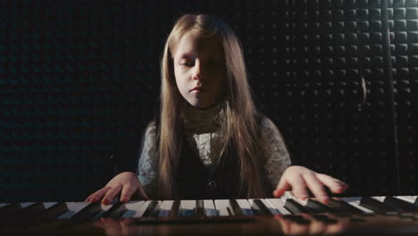 blonde child sits at synthesizer in music studio. little musician in festive dress with digital instrument in sound record center. girl talent development