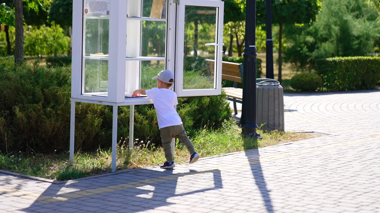 Serious baby boy comes up to a white booth for book crossing in the park. Kid takes a magazine and walks by the alley.