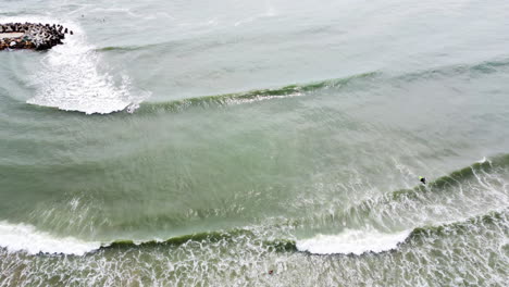 Una-Toma-Vertical-De-Las-Olas-Golpeando-La-Orilla-Del-Mar-Conecta-La-Playa-Mientras-Algunos-Surfistas-Pasan-El-Rato-En-El-Agua