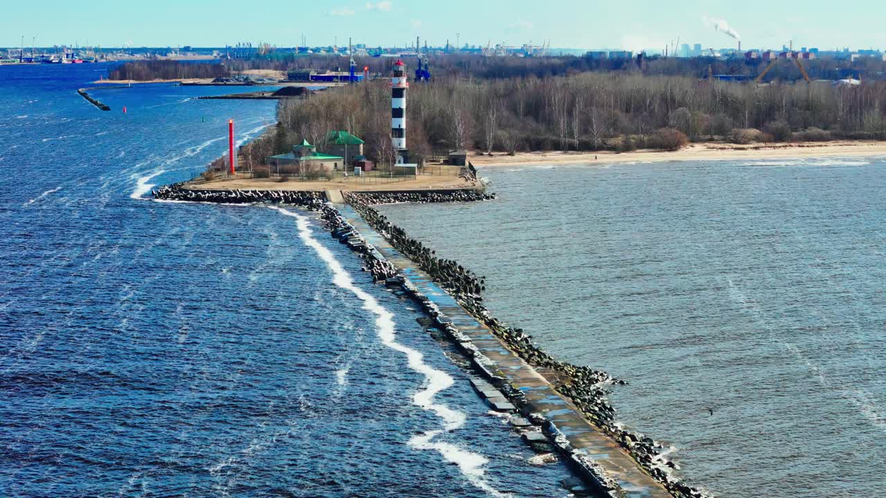 wide-angle Daugavgrīva breakwater and lighthouse in Riga, Latvia, on a bright, clear spring day, separating deep blue river water from the grey-blue Baltic Sea, with a distant industrial shoreline
