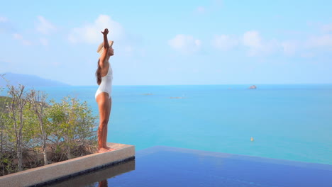 Mujer-Asiática-Libre-De-Pie-En-El-Borde-De-La-Piscina-Infinita-Con-Vistas-Al-Océano-Levanta-Los-Brazos-Hacia-El-Cielo