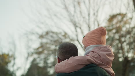 a man in a green jacket is seen carrying his young daughter, who is dressed in a pink jacket and matching beanie, as he points toward the sky