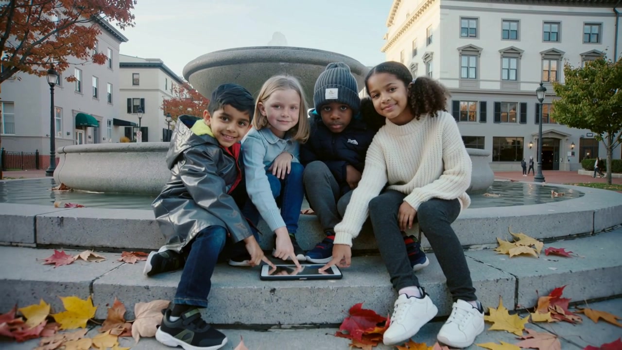 Vier Kinder tippen und wischen auf einem Tablet auf Brunnenstufen, Mäntel tragend, spielend.